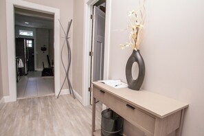 A hallway with a wooden floor, a desk with a vase, and a door with a mirror.