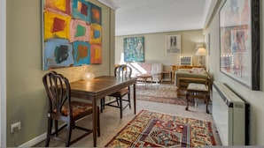 Een eetgedeelte met een houten tafel en stoelen, een kleurrijk abstract schilderij aan de muur, en een kamerbreed tapijt op de vloer.