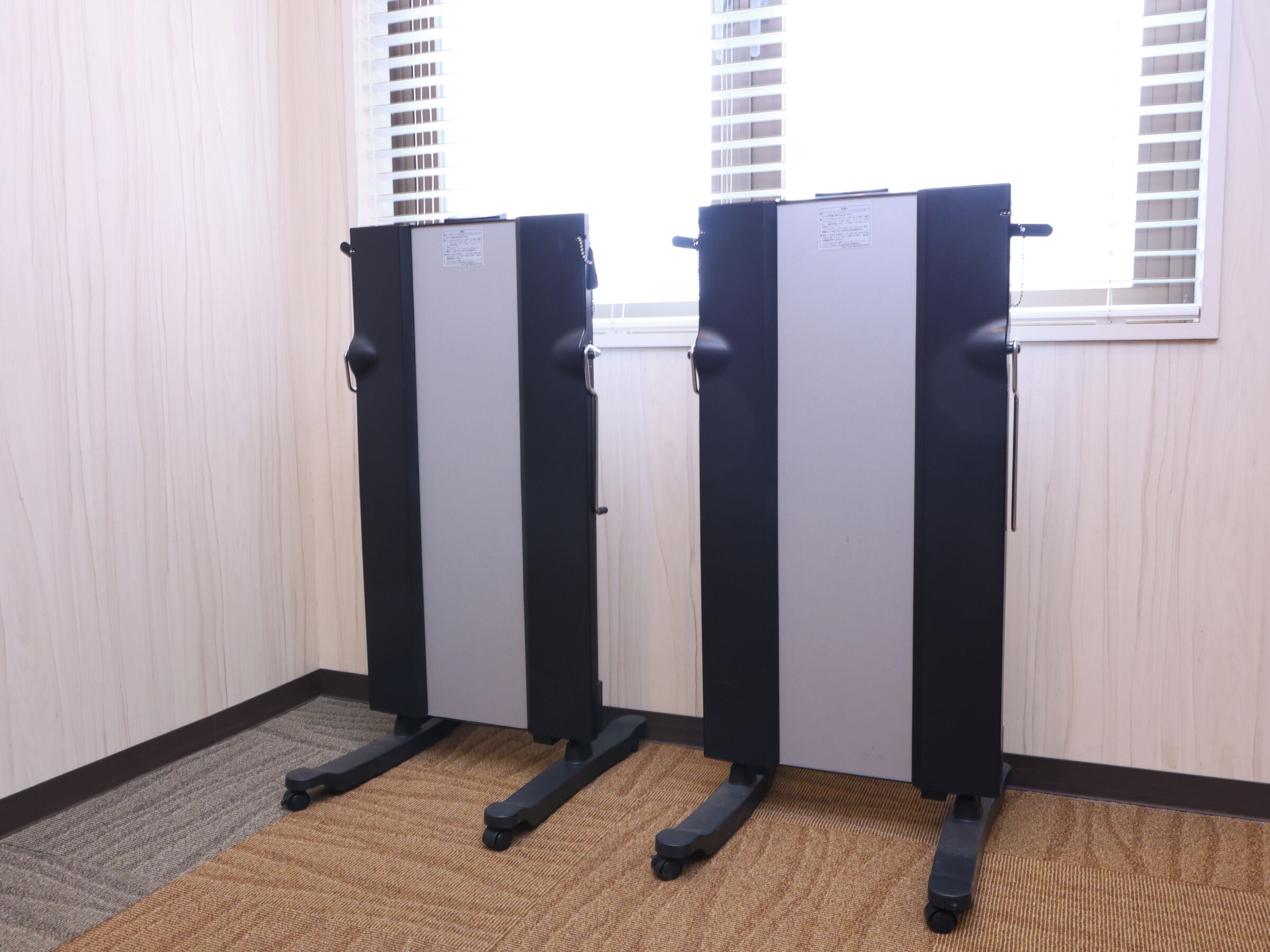 A room with three black and gray exercise machines, a brown carpet, and a window with blinds.