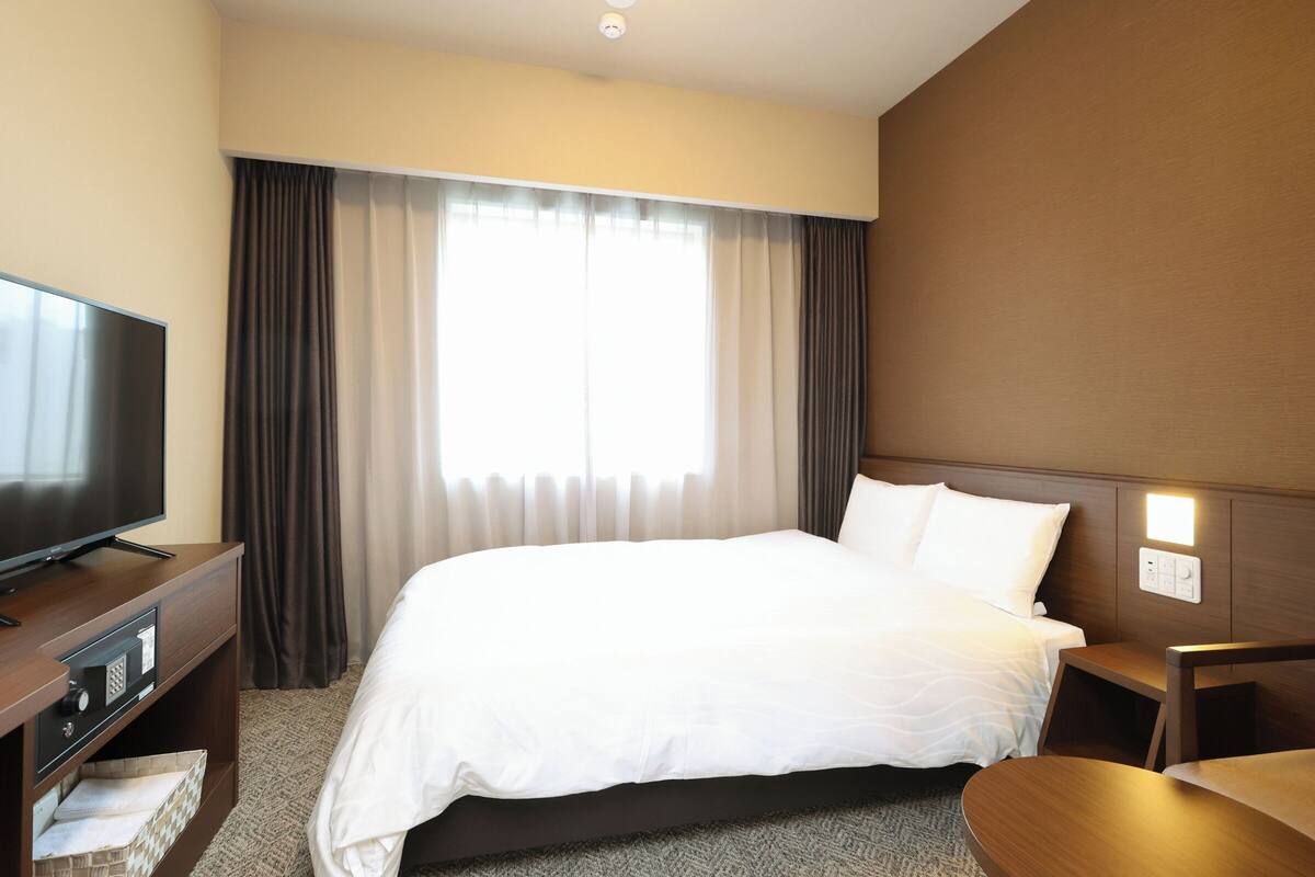 Una habitación de hotel con cama, televisión, escritorio y ventana con cortinas.
