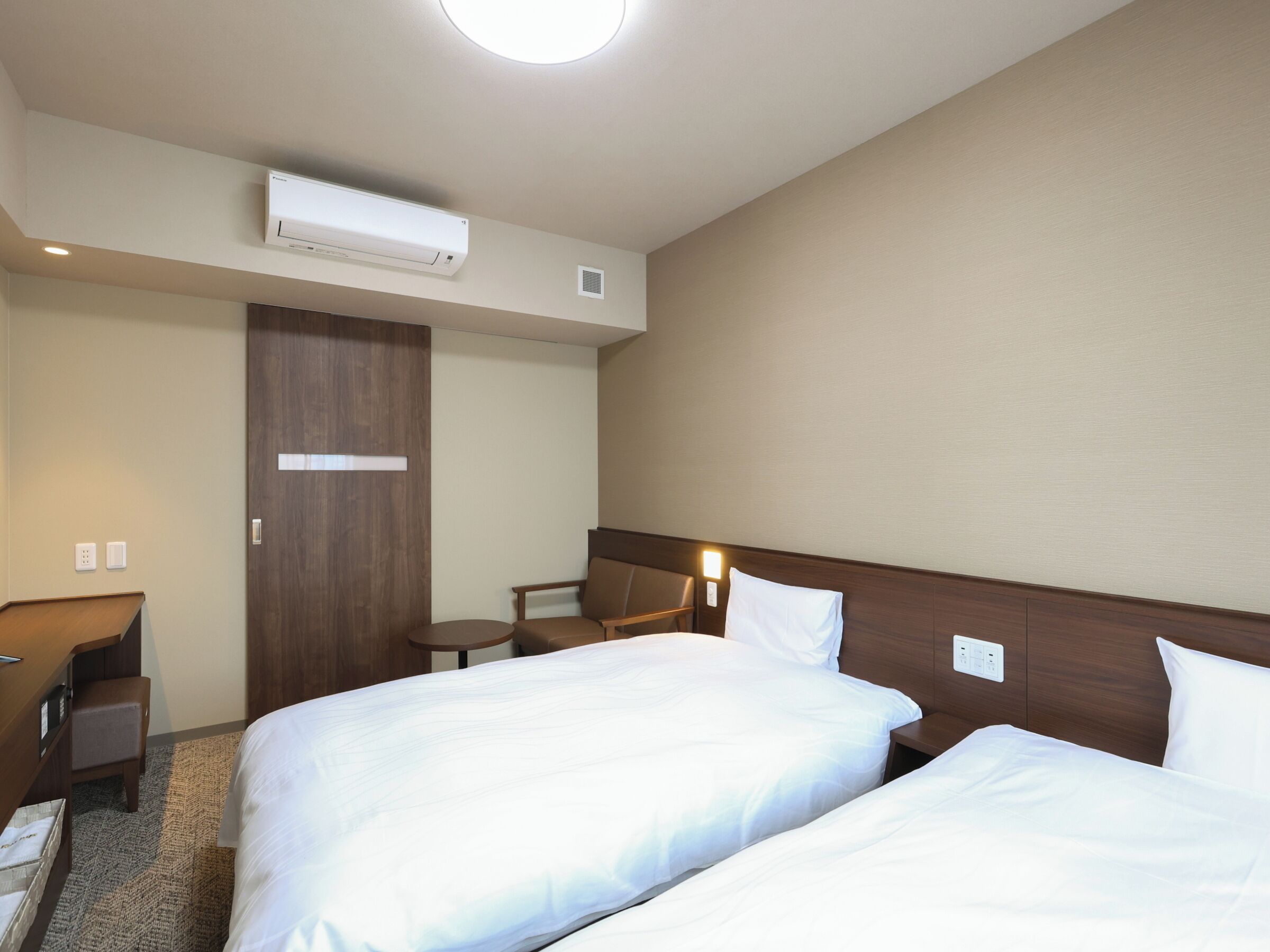 A hotel room with two beds, a desk, a chair, an air conditioning unit, and a door.