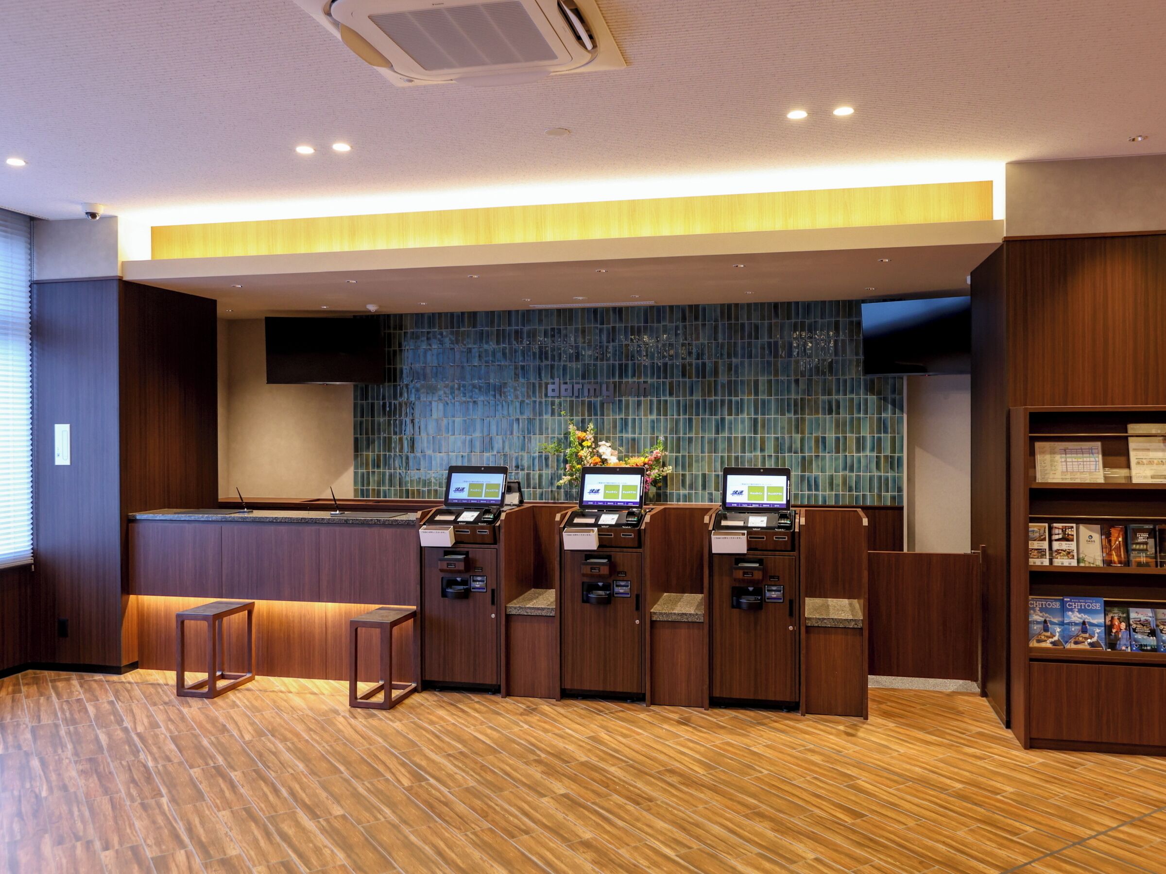 A reception area with a wooden floor, a reception desk, two computer kiosks, a wall with a blue mosaic pattern, and a bookshelf with magazines.