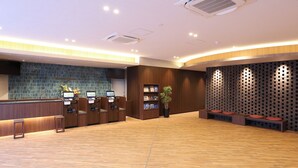 Ein Empfangsbereich mit einem Holzbüro, zwei Selbstbedienungs-Check-in-Terminals, einem Blumentopf und einer Sitzecke mit Kissen.