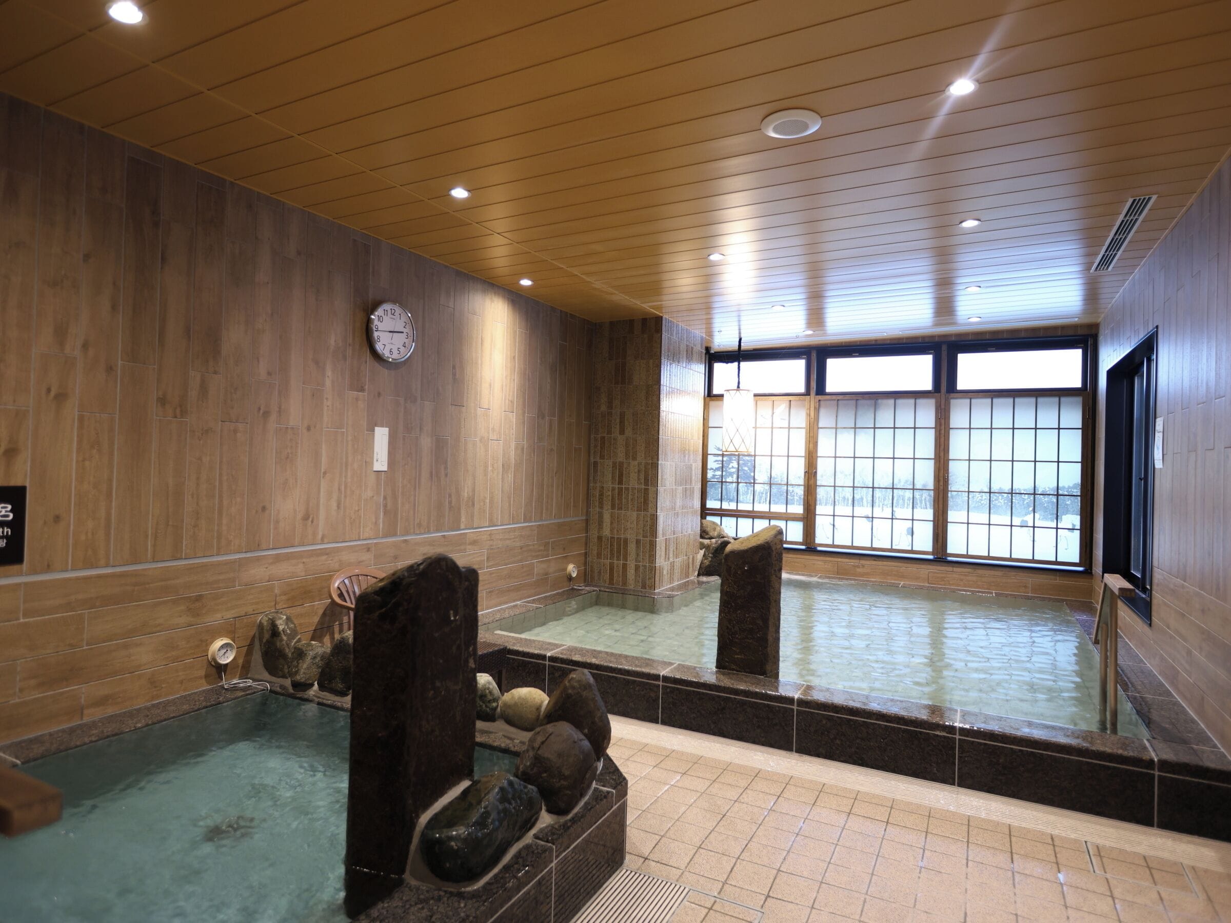 Une salle de spa aux murs en bois, avec une grande fenêtre, une horloge et un jacuzzi agrémenté de rochers.