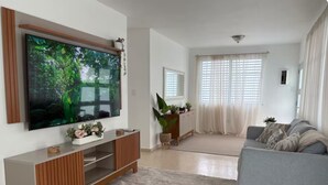 A living room with a flat-screen TV, a grey sofa, a wooden entertainment unit, and a large window with sheer curtains. - Modern 4BR Apartment Near SJU Airport | Fast WiFi • Parking - AC (San Juan)