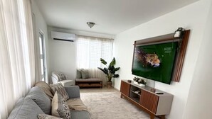 A living room with a grey sofa, a wooden coffee table with a plant, a TV on the wall, and a window with curtains. - Modern 4BR Apartment Near SJU Airport | Fast WiFi • Parking - AC (San Juan)