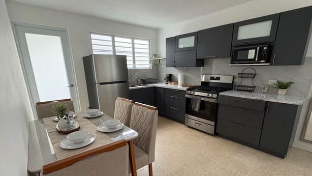 A modern kitchen with dark cabinets, a stainless steel refrigerator, a microwave, a stove, and a dining area with a table set for two. - Modern 4BR Apartment Near SJU Airport | Fast WiFi • Parking - AC (San Juan)