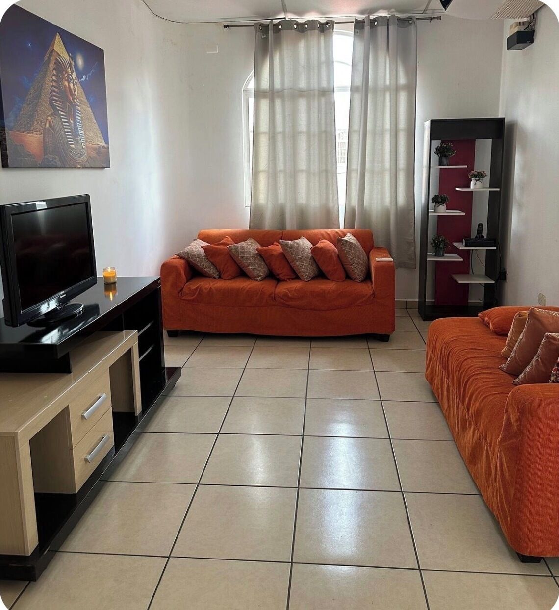 Ein Wohnzimmer mit Fernseher, orangefarbenen Sofas, einem TV-Schrank und einem Wandregal mit Pflanzen.