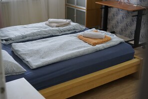 Une chambre avec deux lits, chacun doté d’un matelas bleu et d’une couverture à motifs. On y trouve une table en bois avec une prise électrique et une lampe.