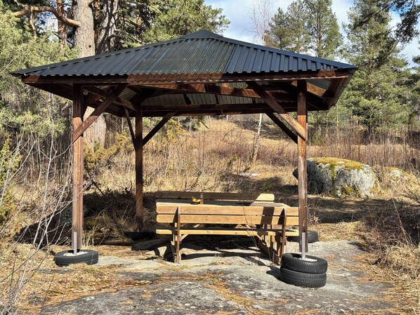 Ein hölzerner Pavillon mit Bank und Tisch, umgeben von Bäumen und Steinen.