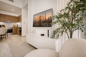 Een moderne woonkamer met een flatscreen televisie aan de muur, een witte salontafel en een eethoek, met een keuken op de achtergrond.