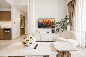 A modern living room with a flat-screen TV, a white sofa, a coffee table with a vase of flowers, and a kitchen in the background. - Luxury 2BR | Full Burj View | Grande Signature (Dubai)