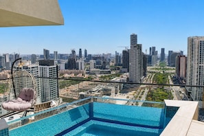 Une piscine à débordement située sur le toit, offrant une vue sur la ville, ainsi qu’un fauteuil suspendu et un ciel dégagé.