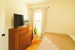 Ein Schlafzimmer mit einem hölzernen Schminktisch, einem Fernseher darauf, einem Bett mit beiger Bettdecke, einer Topfpflanze und einem Fenster mit Vorhängen.