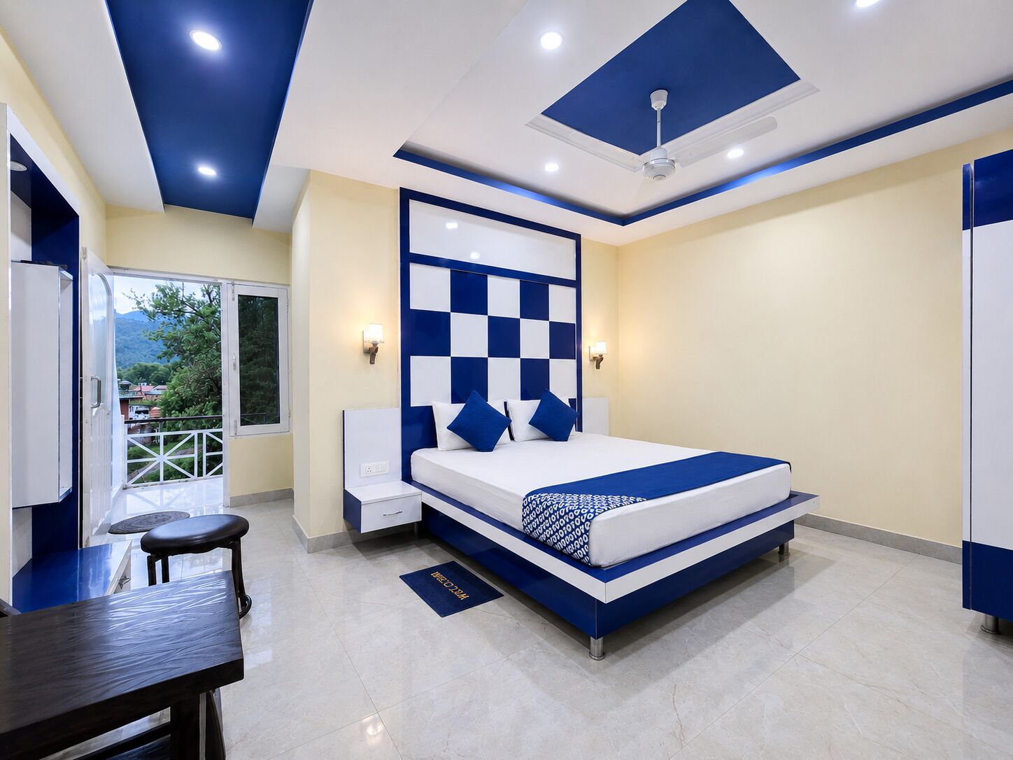 A modern bedroom with a checkered headboard, a bed with blue and white bedding, a desk with a chair, and a balcony with a view.