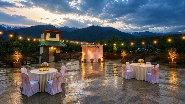 An outdoor wedding setup with tables and chairs, a stage, and string lights against a mountainous backdrop. - Osho Hotel, Restaurant & Banquet (Palampur)