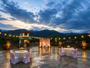 Montaje para una boda al aire libre con mesas y sillas, un escenario y luces colgantes con montañas al fondo.