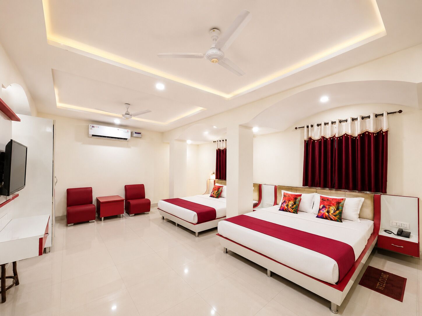 A hotel room with a large bed, two smaller beds, a desk, a TV, and a ceiling fan.