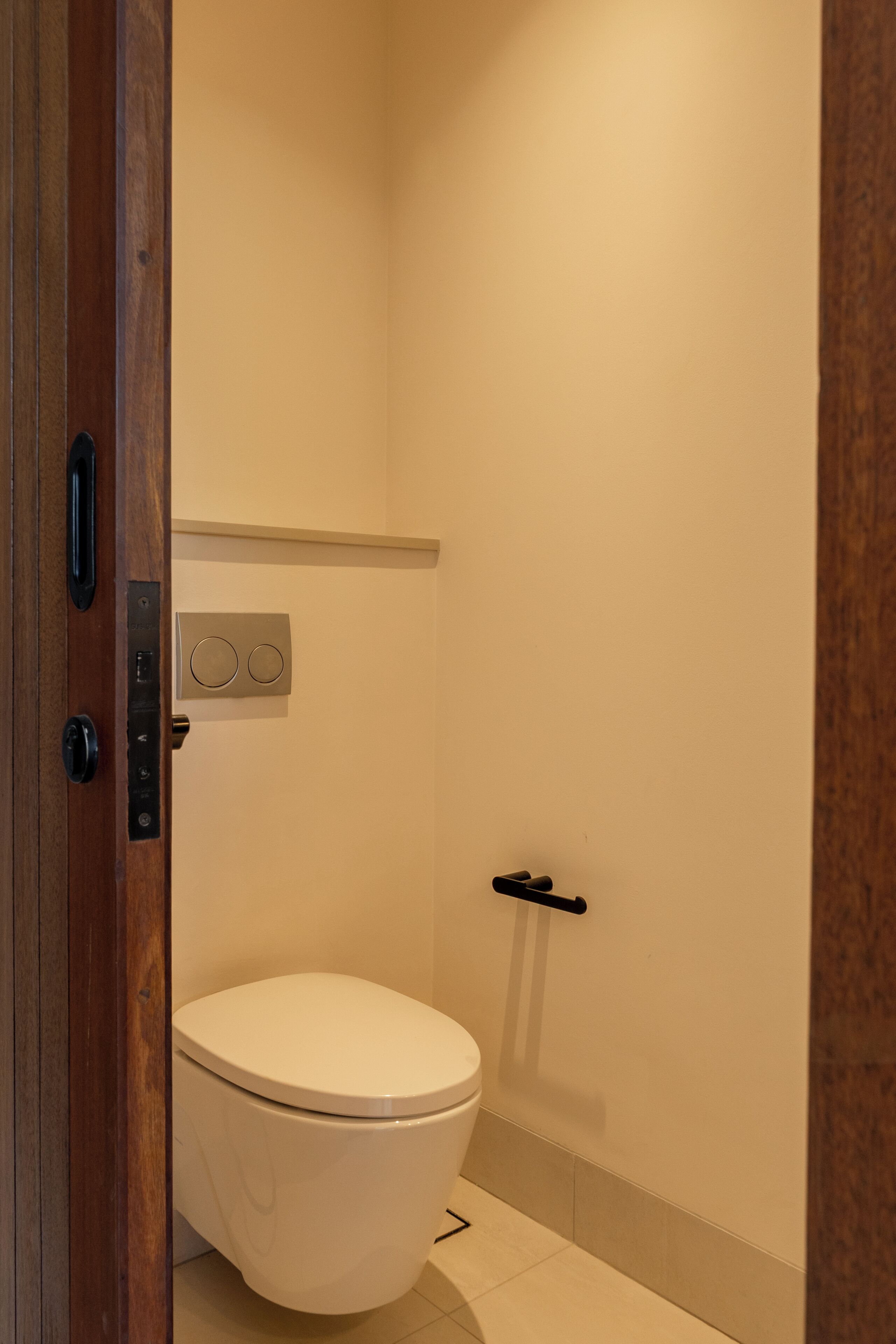 Un baño con un inodoro blanco, un accionador de descarga en la pared y una puerta con manija.