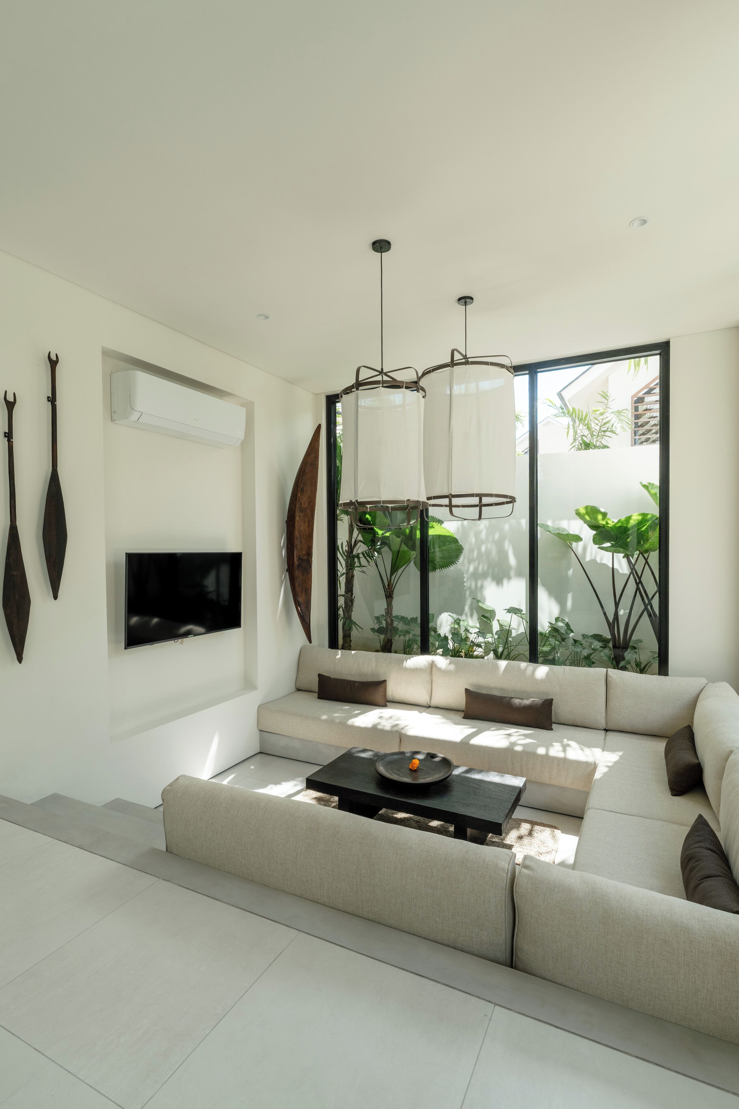 A modern living room with a sofa, a flat-screen TV, a coffee table, and a large window with plants.