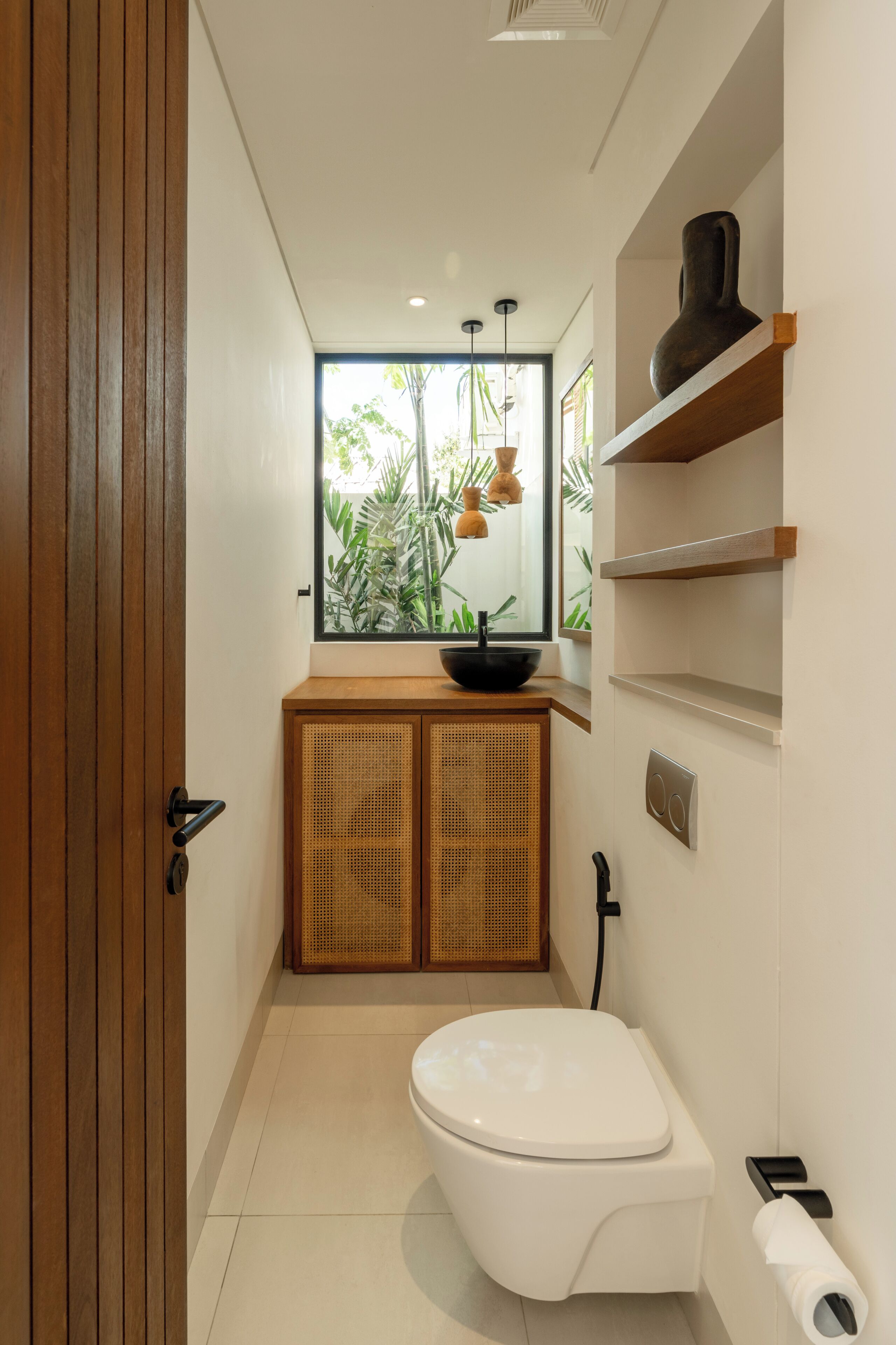 A modern bathroom with a toilet, a sink with a black basin, a window with plants, and wooden shelves.