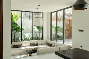 A modern living room with a large sofa, a coffee table, and a dining table. There are large windows with a view of greenery outside. - Aluka Bingin (Pecatu)