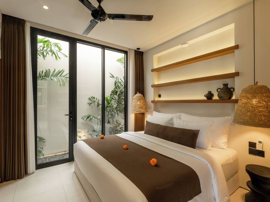 Een moderne hotelkamer met een groot bed, een plafondventilator, een schuifdeur naar een balkon en een wandplank met decoratieve objecten.