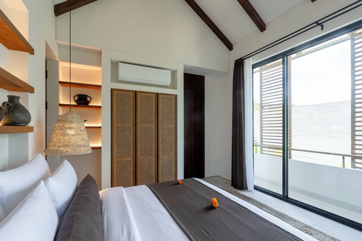Une chambre à coucher moderne avec un lit, une lampe suspendue, une étagère avec un vase et une grande fenêtre avec des stores.