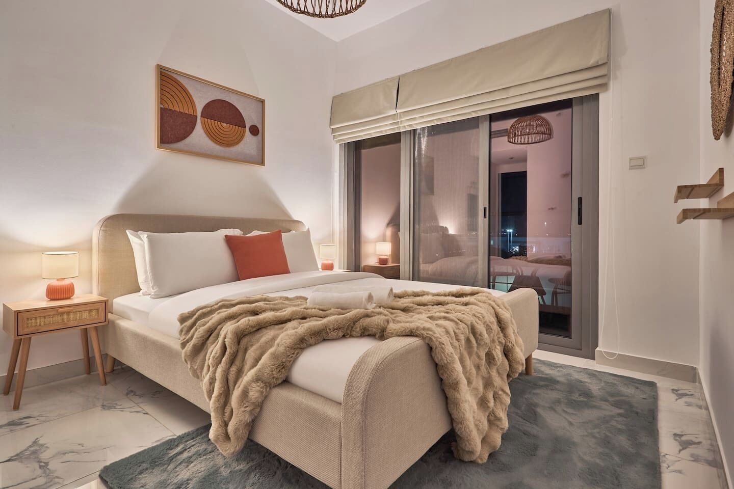 A bedroom with a large bed, a nightstand, a lamp, a window with a view, and a painting on the wall.