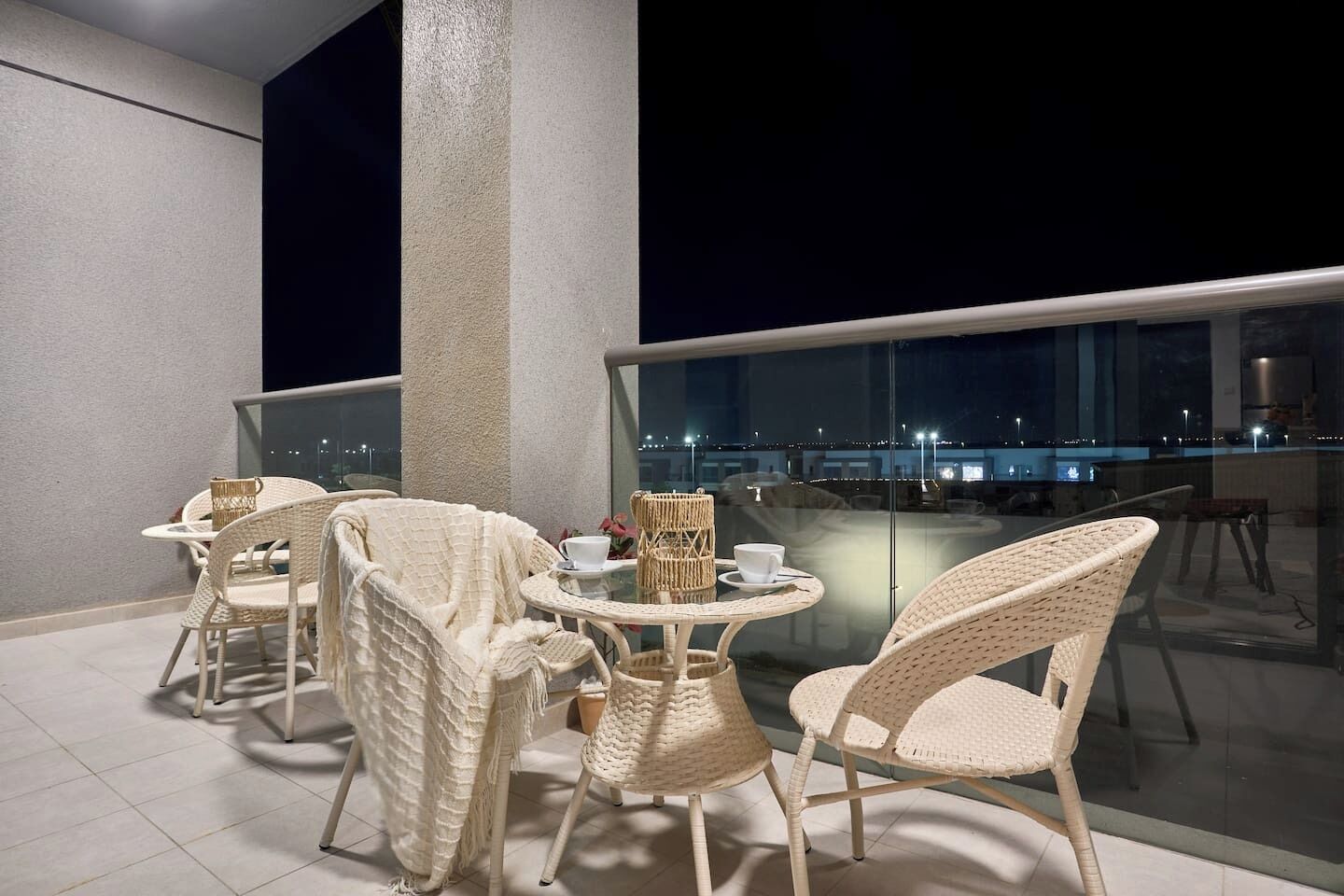 A balcony with wicker furniture, a glass table, and a view of the city at night.