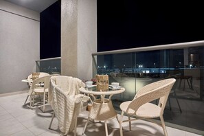 Un balcon agrémenté de meubles en osier, d’une table en verre et offrant une vue sur la ville la nuit.