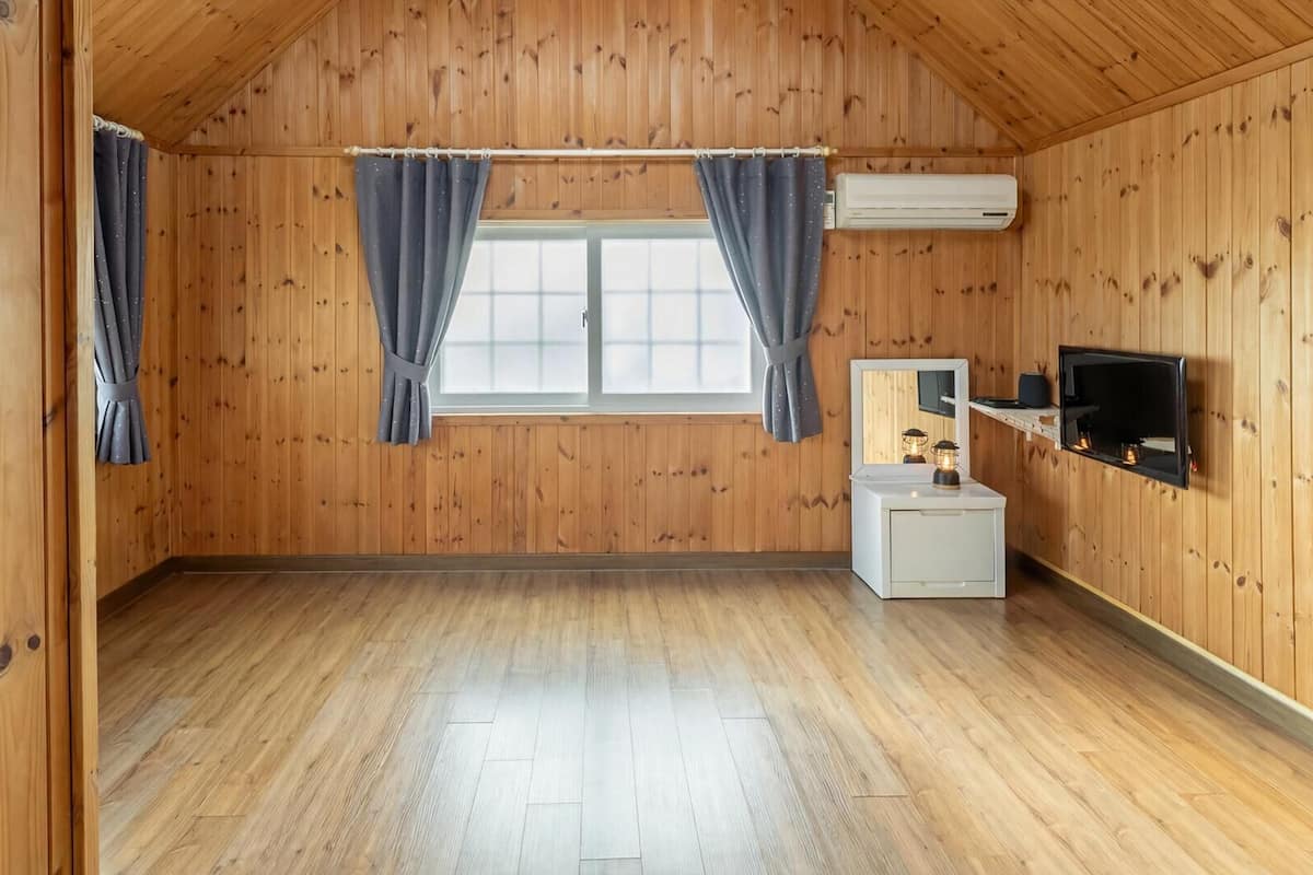 Una habitación con paredes de madera, una ventana con cortinas, un televisor de pantalla plana y una mesita de noche con lámpara.