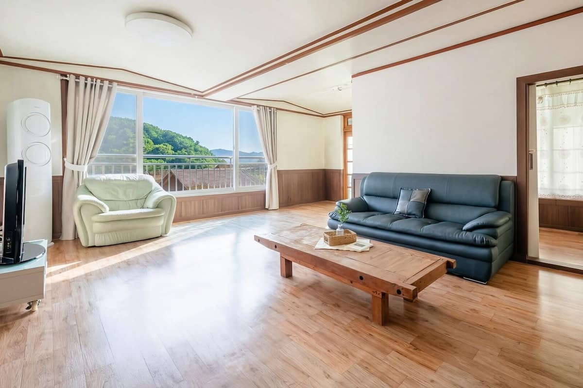 Un salón amplio con piso de madera, un sofá, una butaca, una mesa de centro y ventanas grandes que dan a vistas de vegetación.