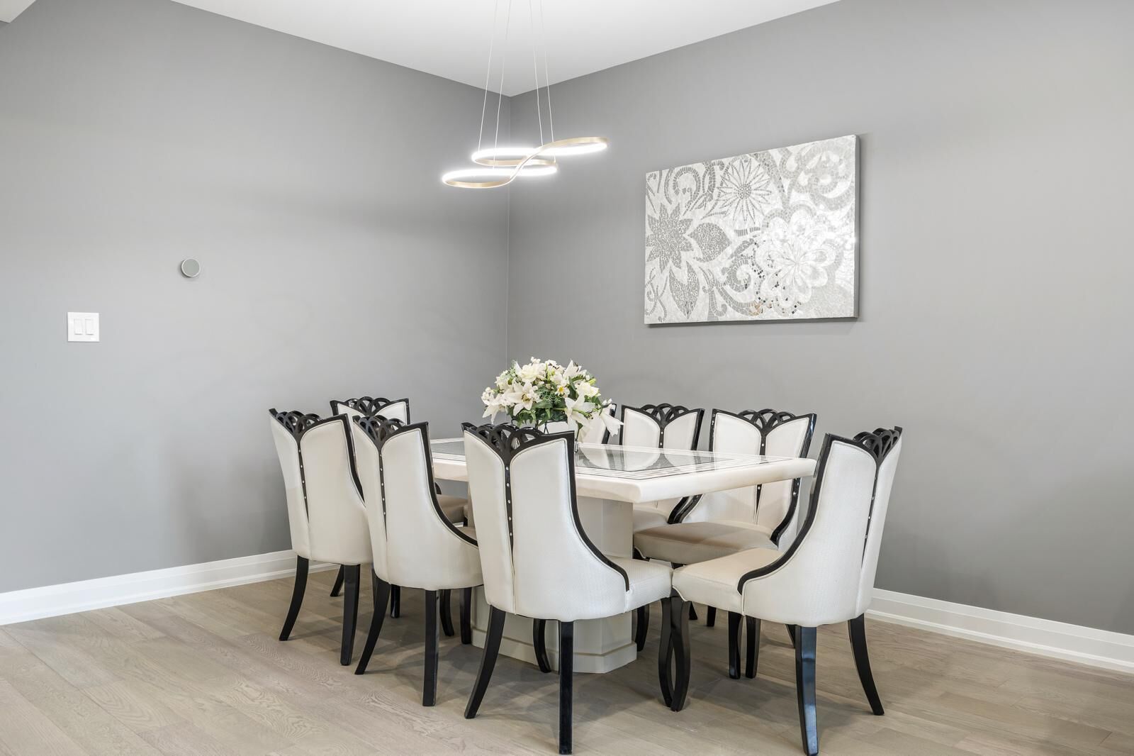A dining room with a round table, six chairs, a vase of flowers, and a wall-mounted artwork.