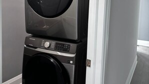 Uma máquina de lavar e secar empilhadas em um corredor, com uma parede cinza e uma porta à direita.
