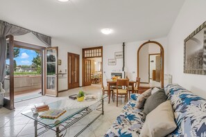 Ein Wohnzimmer mit einer Couch, einem Glastisch, einem Essbereich mit Tisch und Stühlen und Blick durch eine offene Tür.