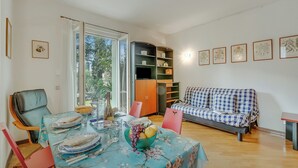 Un comedor con una mesa dispuesta para cuatro personas, un sofá, una estantería y una ventana con vistas a zonas verdes.
