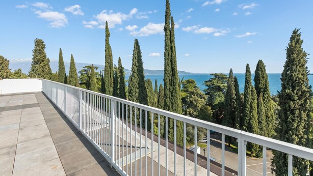 Ein Balkon mit weißer Brüstung, der auf eine Küstenlandschaft mit hohen Bäumen und blauem Himmel blickt.