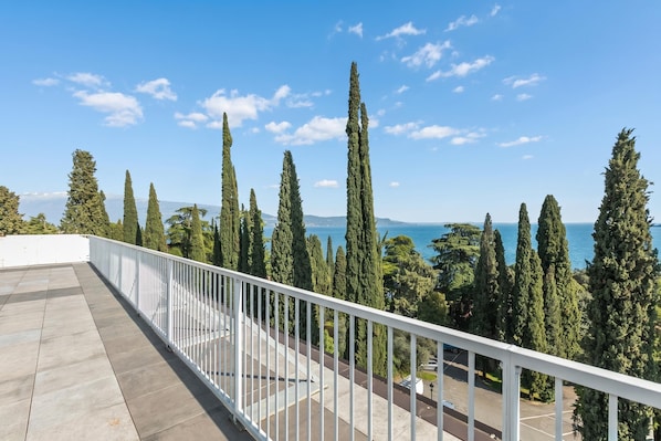 Ein Balkon mit weißer Brüstung, der auf eine Küstenlandschaft mit hohen Bäumen und blauem Himmel blickt.