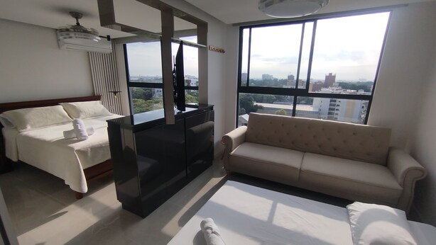 A modern hotel room with a bed, a sofa, a TV, and a large window offering a city view. - Loft Deluxe 12th Floor Cali Velodrome View (Cali)