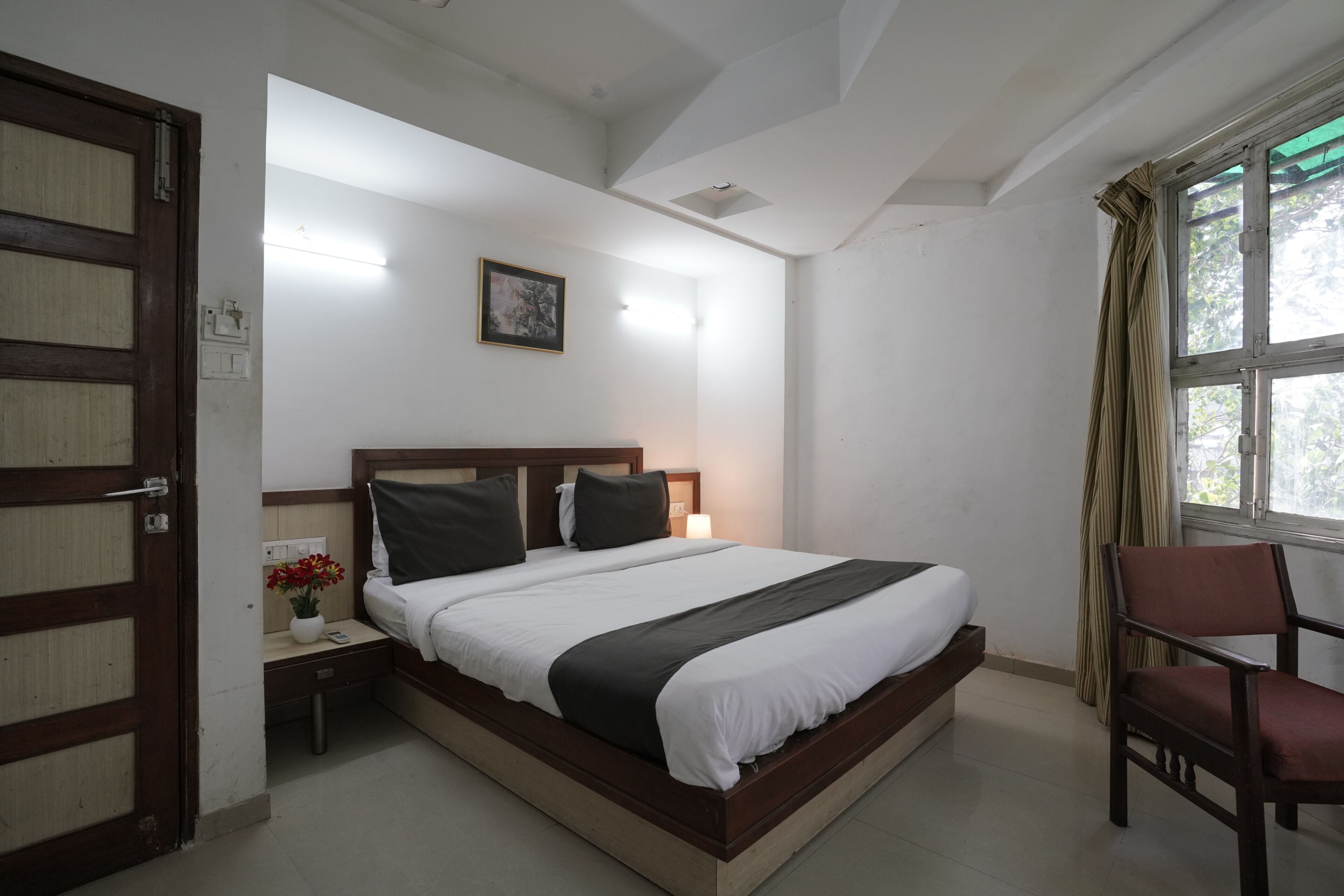 A hotel room with a bed, a chair, a nightstand, a wall-mounted light, a window with curtains, and a ceiling light.