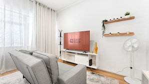Un soggiorno moderno con un divano grigio, una TV a schermo piatto, un mobile TV bianco, una ventola a piantana e una finestra con tende leggere.