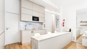 Una cucina moderna con un piano di lavoro in marmo, un forno a microonde e un lavandino. Sono presenti armadietti bianchi, un estintore e una piccola pianta sul bancone.