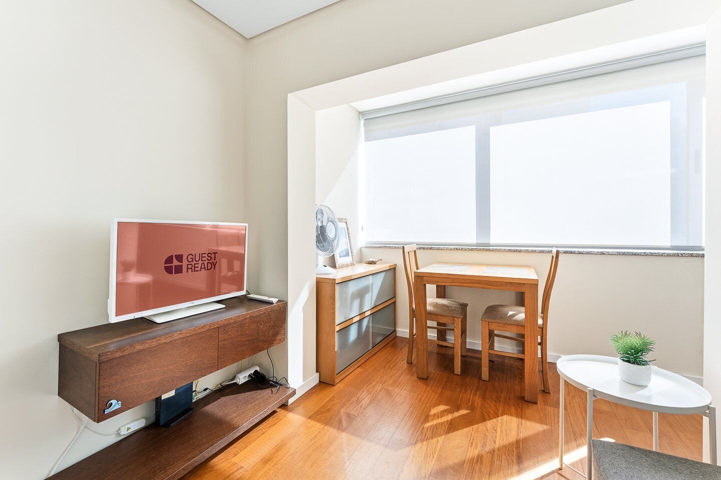 Une chambre d’hôtel équipée d’une télévision affichant le message « CHAMBRE PRÊTE POUR L’ARRIVÉE DES CLIENTS », d’un bureau en bois avec deux chaises, d’une petite table avec une plante et d’une fenêtre avec des stores.