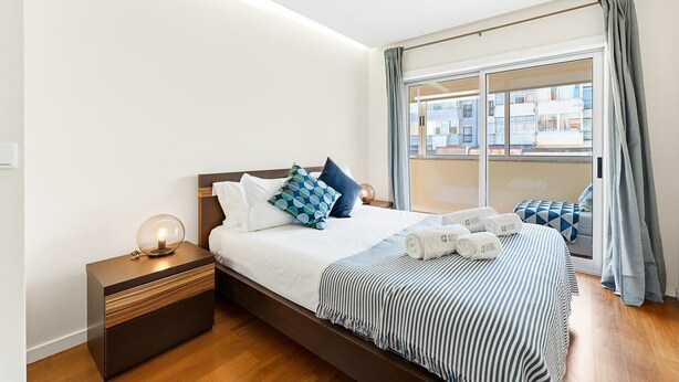 Una habitación de hotel moderna con una cama grande, una mesita de noche con lámpara, vista a edificios por la ventana y un balcón con vistas.
