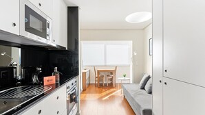 Una cocina compacta con microondas, cafetera y horno. Hay una zona de comedor con una mesa y sillas, y un sofá gris.