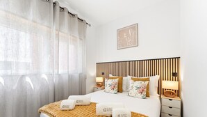 A hotel room with a bed, a bedside table, a nightstand, a lamp, a window with curtains, and a wall-mounted picture. - GuestReady - Apartment near beach retreat (Vila do Conde)