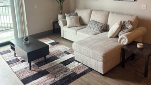 Un salon moderne avec un canapé d’angle, une table basse et un tapis. On y trouve une plante en pot et une œuvre d’art abstraite accrochée au mur.