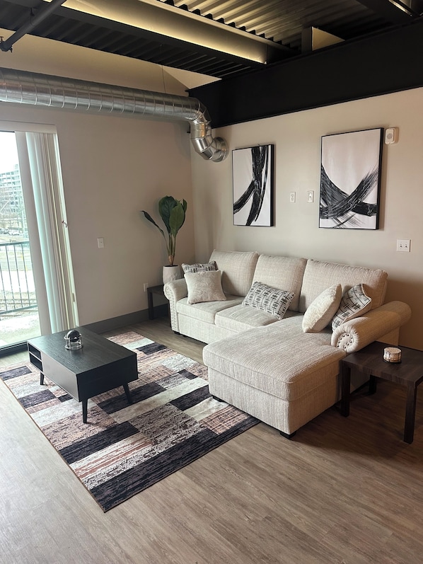 A modern living room with a sectional sofa, a coffee table, and a rug. There is a potted plant and abstract art on the wall.