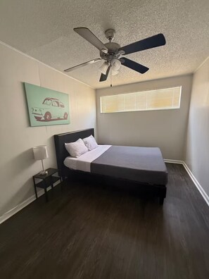 Um quarto com cama, ventilador de teto, janela com persianas, mesa de cabeceira com abajur e uma parede com ilustração de carro.